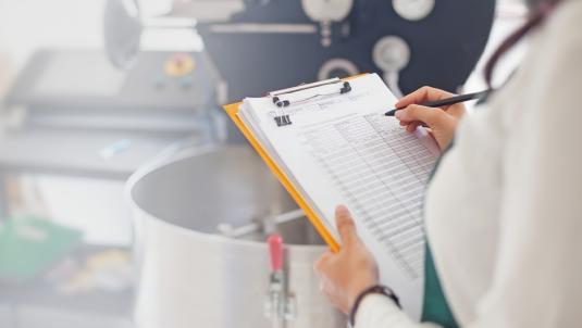A person writing on a clipboard in front of a steel pot.