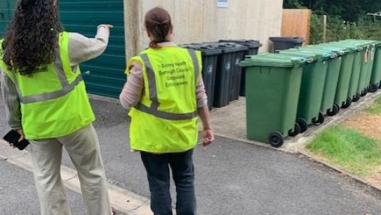 2 people in hi vis in front of a row of wheelie bins