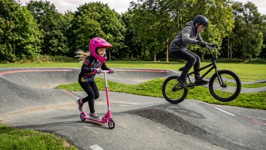 Two children riding a bike and a scooter on a pump track