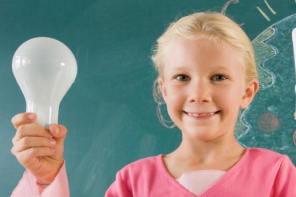 [A child holding up an old fashioned lightbulb and an energy efficient lightbulb. They are standing in front of a blackboard with a drawing of Earth on it]