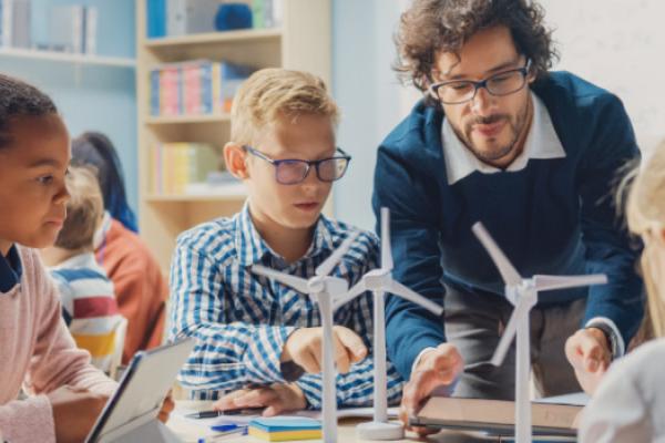 [A teacher and a group of children in a classroom looking a model wind turbines]