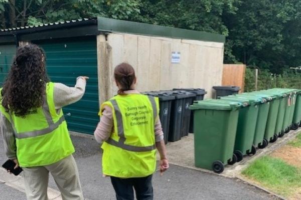 2 people in hi vis in front of a row of wheelie bins