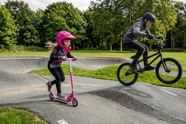 Two children riding a bike and a scooter on a pump track
