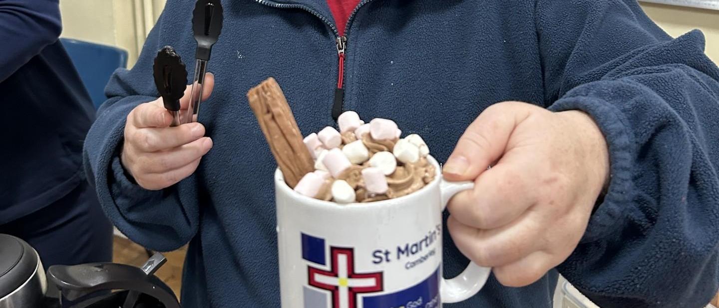 church volunteer holding a large hot chocolate with cream, marshmallows and a flake on top