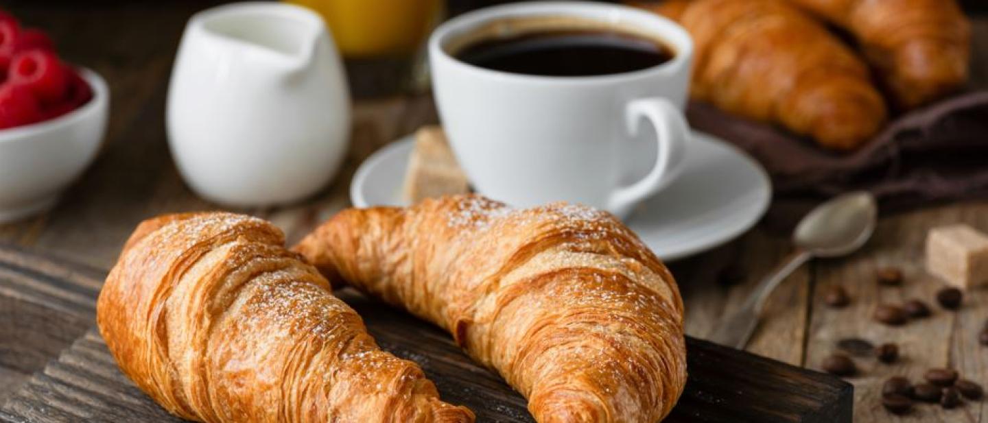two croissants on a wooden board with a black coffee in a white tea cup on a saucer with a white milk jug adjacent to it