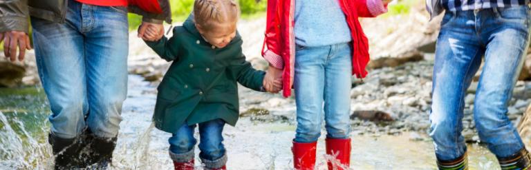 [A family holding hands and wearing wellington boots in a stream]