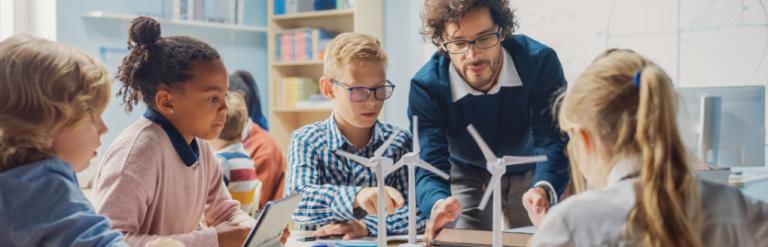 [A teacher and a group of children in a classroom looking a model wind turbines]
