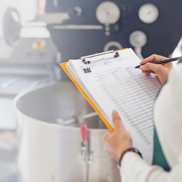 A person writing on a clipboard in front of a steel pot.