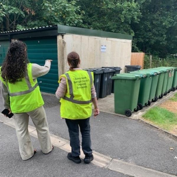 2 people in hi vis in front of a row of wheelie bins