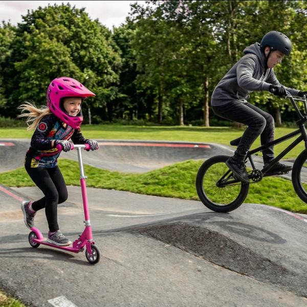 Two children riding a bike and a scooter on a pump track