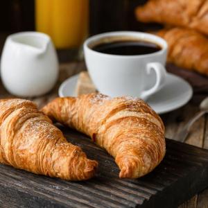 two croissants on a wooden board with a black coffee in a white tea cup on a saucer with a white milk jug adjacent to it